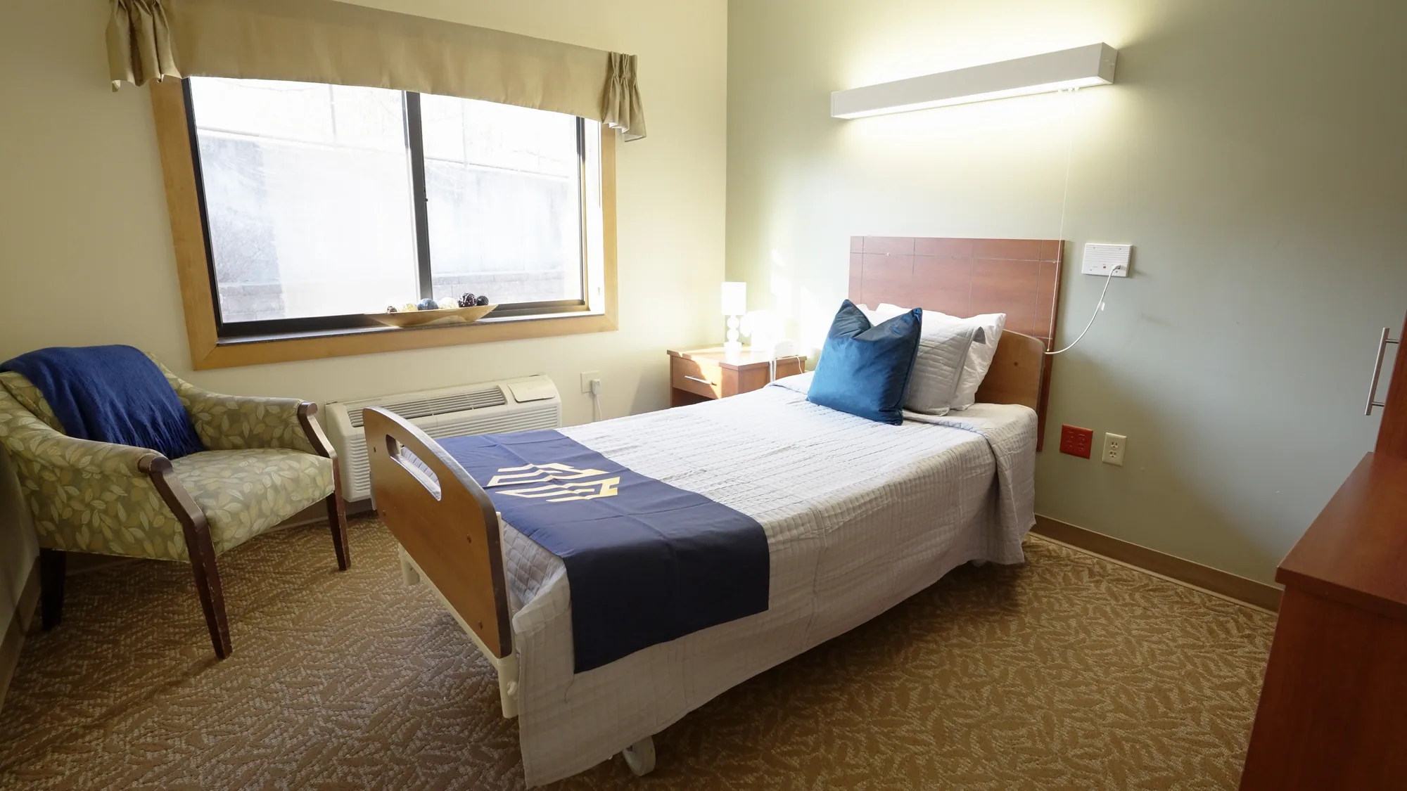 A resident room with a made bed, bedside lamp and armchair