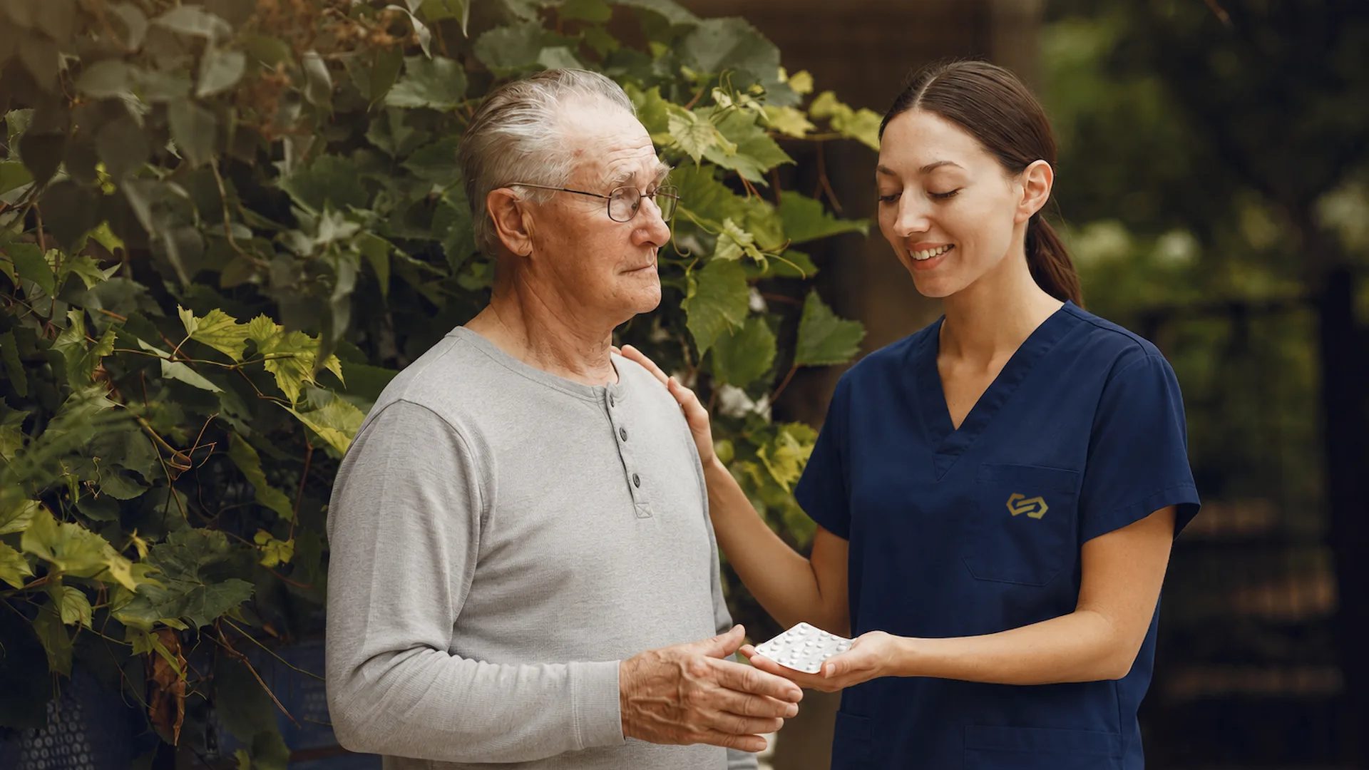 A resident and a nurse walking together outdoors among greenery